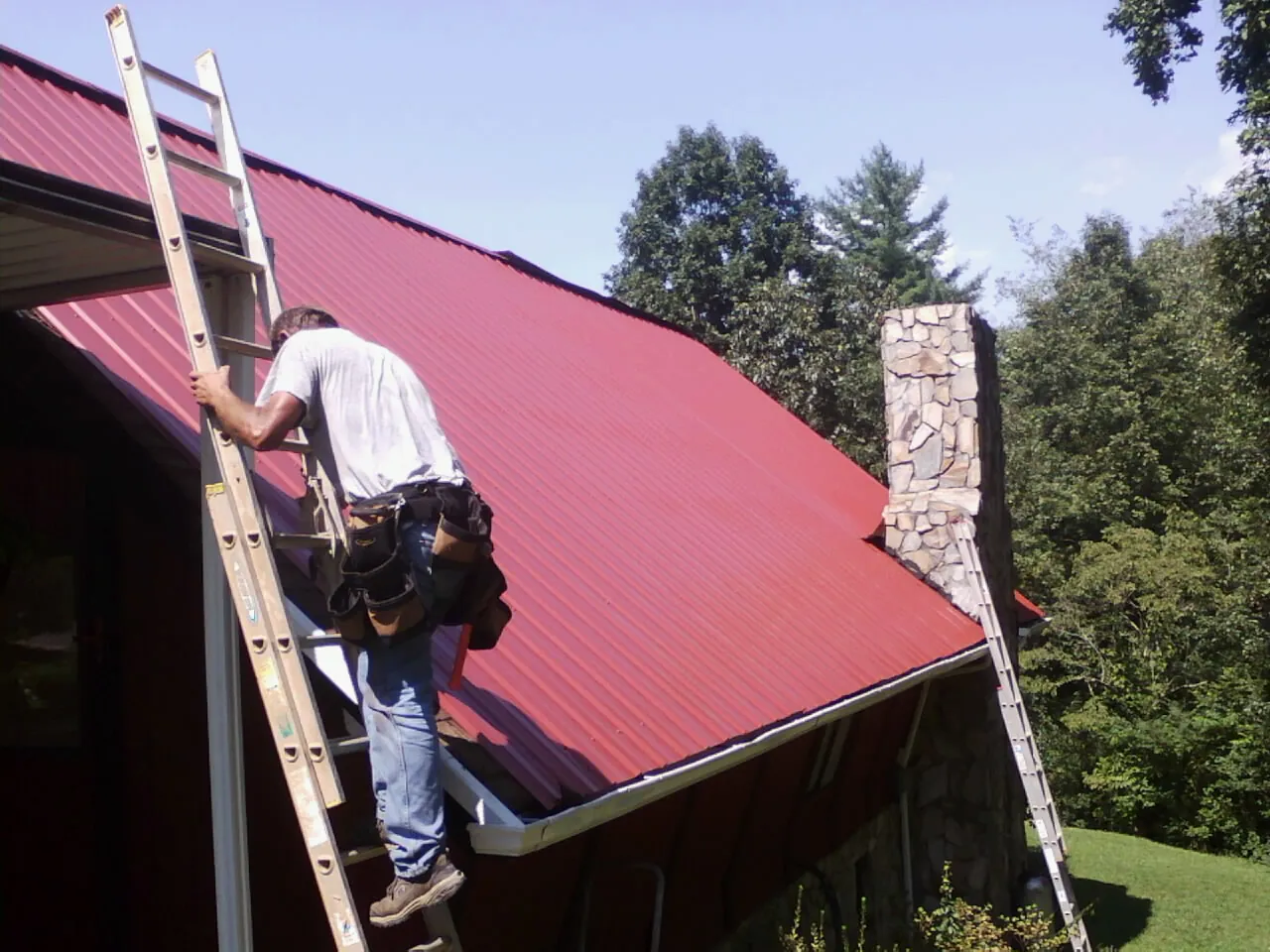 Roofer on a ladder beside a red metal roof and stone chimney — Soffit & Fascia Repair by Roofing Brothers in Fayetteville
